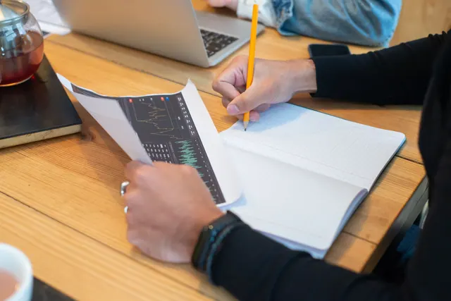 Close-up of hands writing notes on notebook during business meeting with laptop and charts. Content Marketing Analytics Guide for Teams